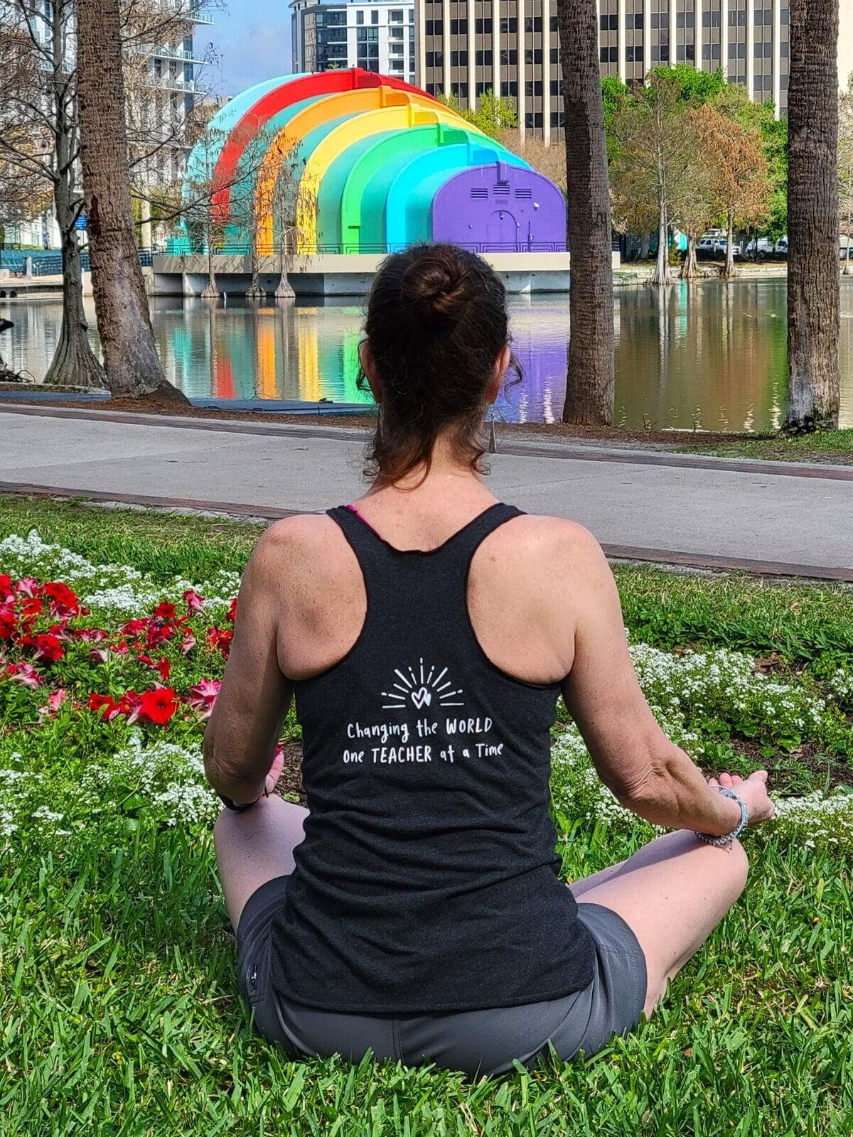 Back view of Sheila in a seated yoga pose, wearing a shirt that reads 'Changing the world one teacher at a time,' with a rainbow stage backdrop